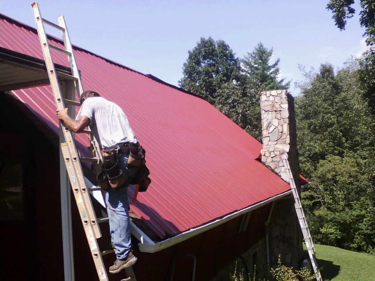 Roofer on a ladder beside a red metal roof and stone chimney — Soffit & Fascia Repair by Roofing Brothers in Coolin
